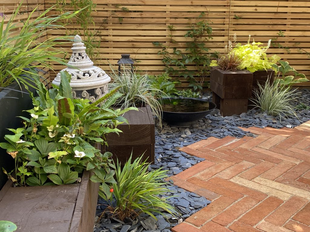 A tranquil patio design with a water feature, lush greenery, and wooden planters on slate stones.