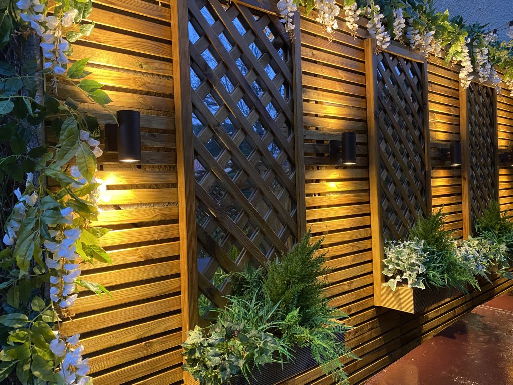 Wooden trellis wall illuminated with modern lighting and adorned with greenery.